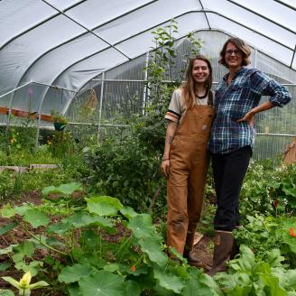 Two women stand inside a seasonal high tunnel surrounded by green crops growing in the ground.