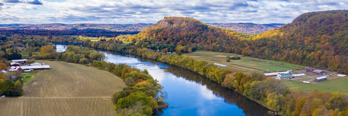 Aerial view of a river with trees on both sides running through agricultural land. There are mountains with fall foliage in the background and cloudy blue skies above.