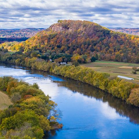 Aerial view of a river with trees on both sides running through agricultural land. There are mountains with fall foliage in the background and cloudy blue skies above.