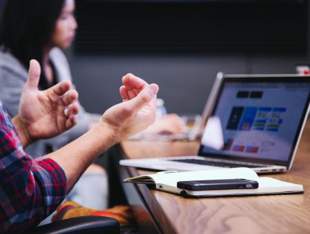 Caucasian hands in front of laptop, woman sits in background at a meeting